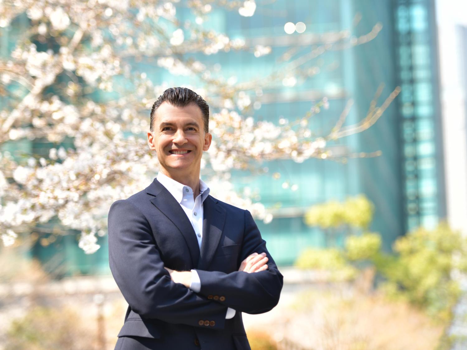 A man in a business suit, with his arms crossed, smiling for the camera, outside on a beautiful, sunny day