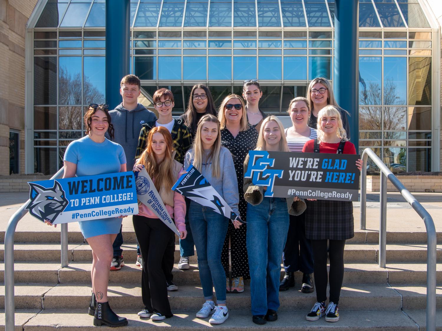 A group of students pose for a photo on the steps of a building. It's a sunny day.
