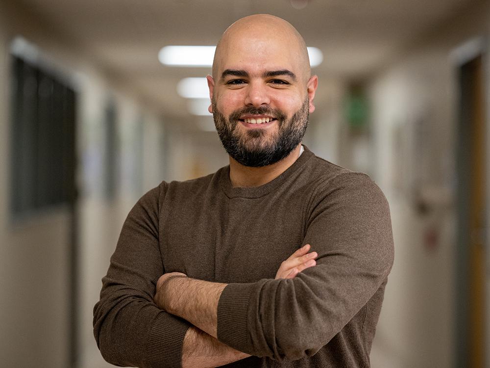 Smiling bald man with dark beard and mustache and wearing brown long-sleeve t-shirt stand with arms crossed in classroom building.  
