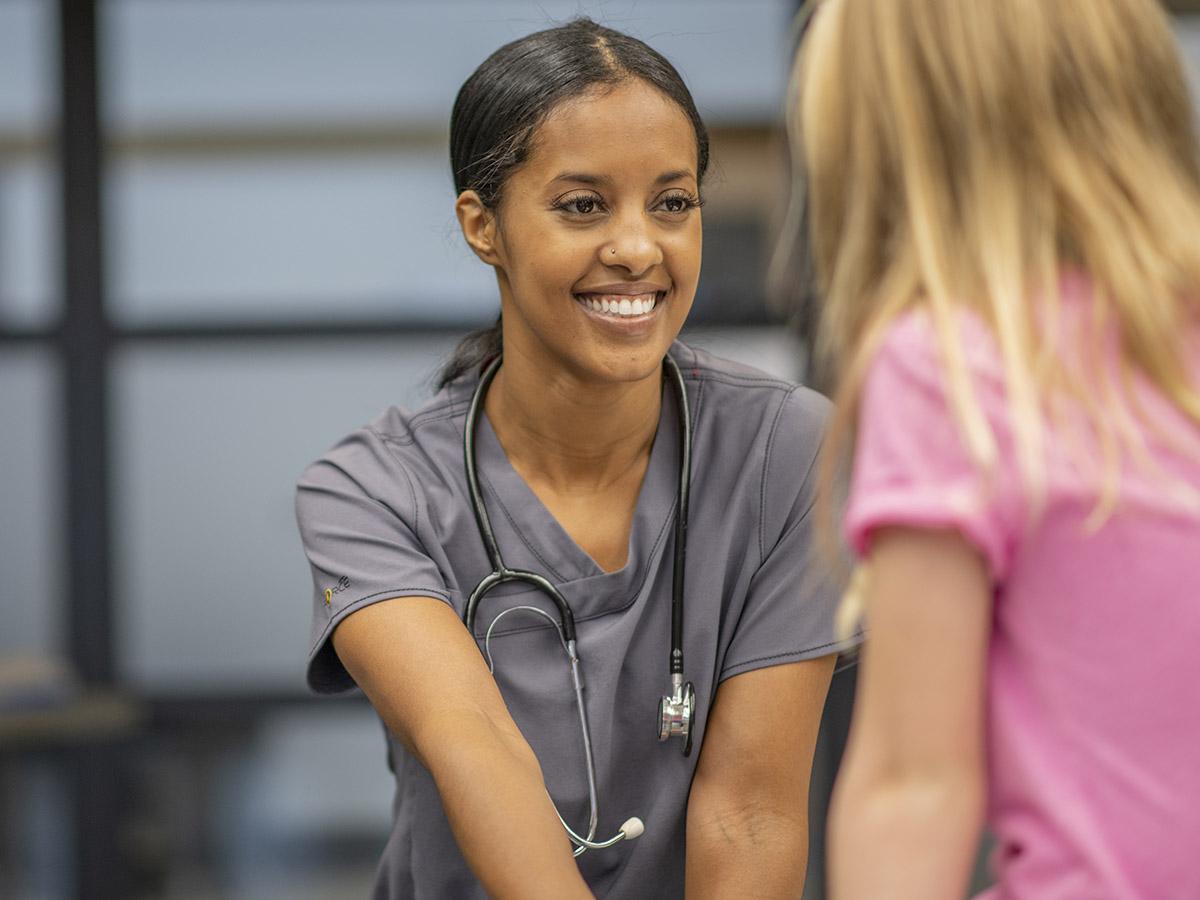 A school nurse wearing scrubs and a stethoscope assists a young student.