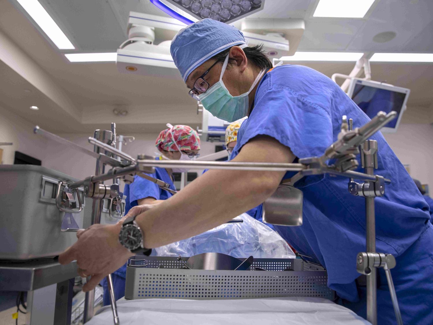 Dr. Johnny Hong, wearing surgical scrubs, works with a piece of equipment in the foreground of an operating room. Other staff members are working in the background.
