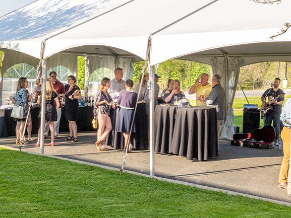 People gather under a white tent on a green lawn on a sunny evening. There are high-top tables under the tent.