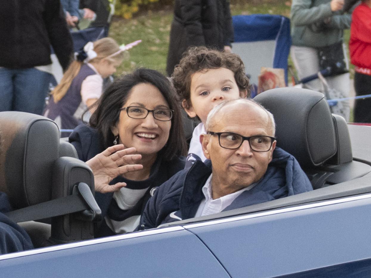 President Neeli Bendapudi sits with her family in an open-topped car, along a parade route. Bendapudi is waving at the camera.