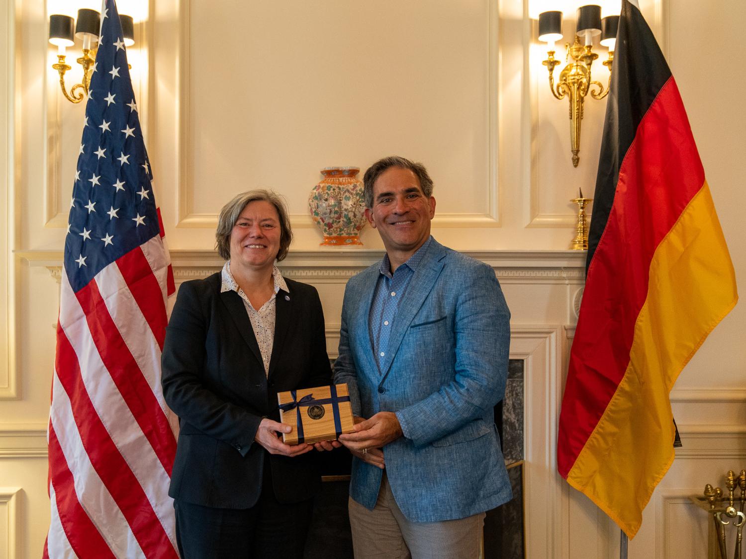 A man presenting a boxed gift to a woman, flanked by the American and German flags
