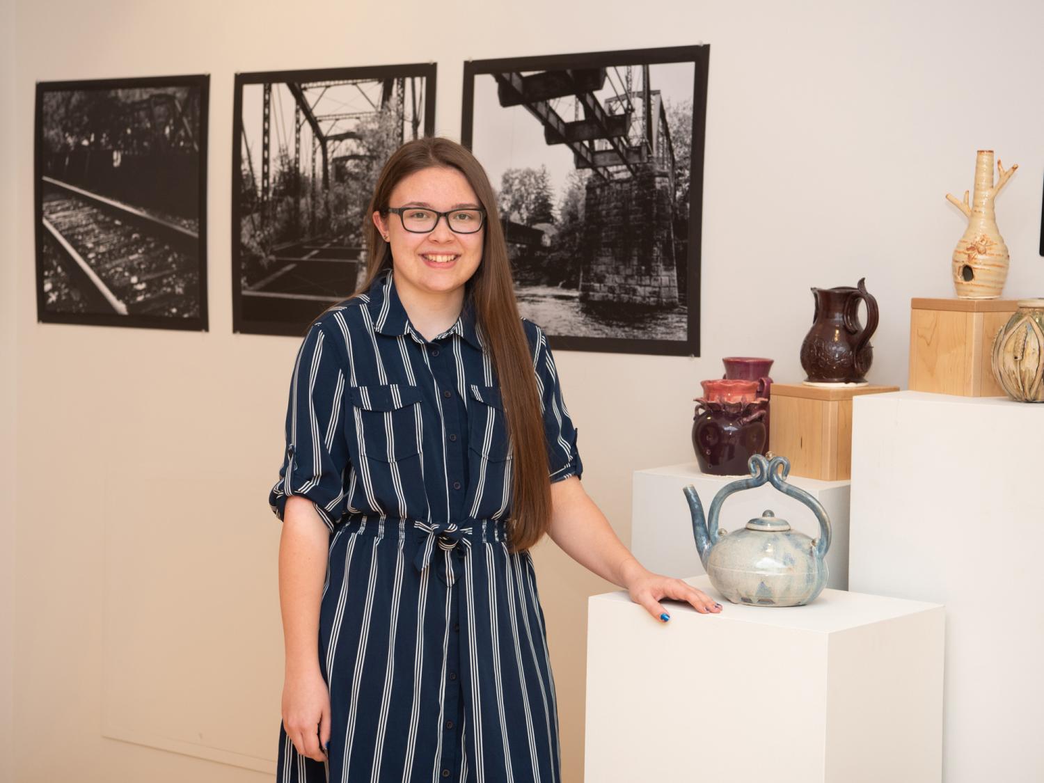 Alexis Burrell standing in The Gallery at Penn College with photographs behind her and ceramics beside her on stands.