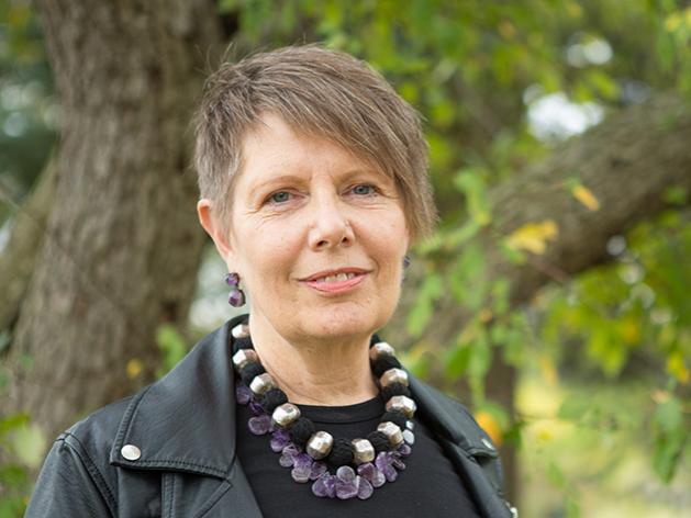 Robin Bower is pictured outdoors with trees in the back ground. She is wearing a black shirt, a chunky necklace and has short hair with side-swept bangs.