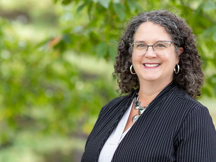 A headshot of Jenifer Cushman standing outdoor in front of a wooded area. She is wearing a dark blazer and white shirt. 