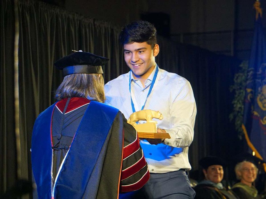 Male student in white dress shirt receiving a lion statue on a stage.