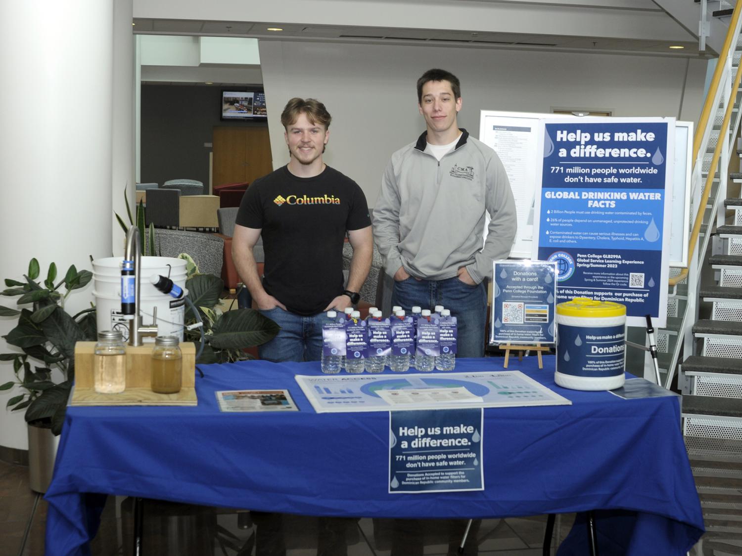 Two people staffing a table covered with information. A poster stands behind them that illustrates information about the need for safe drinking water.