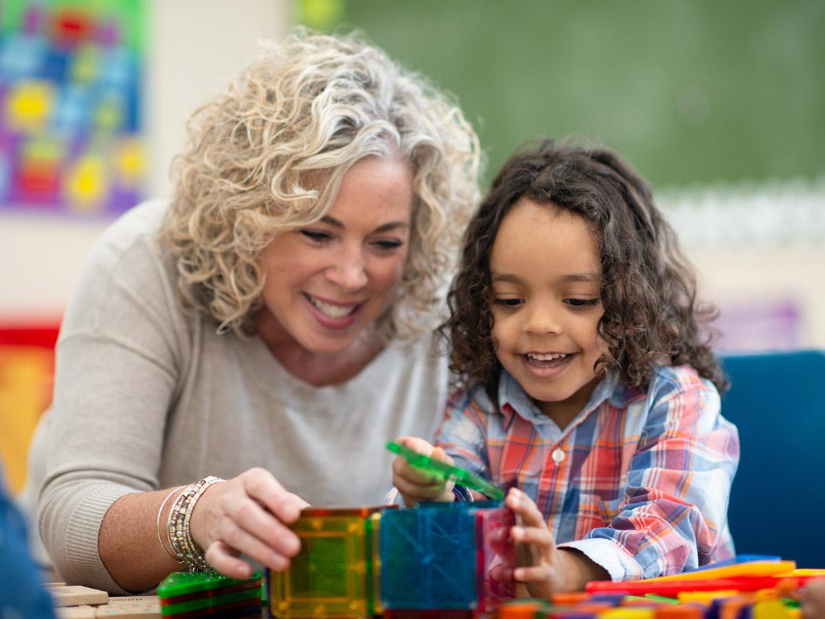 A teacher and child play with blocks in a classroom.