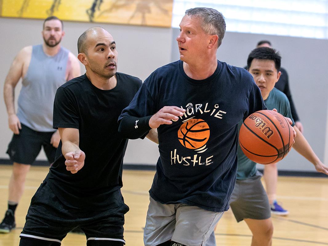 Man wearing athletic clothing dribbles basketball up a basketball court as other men chase him and look on.