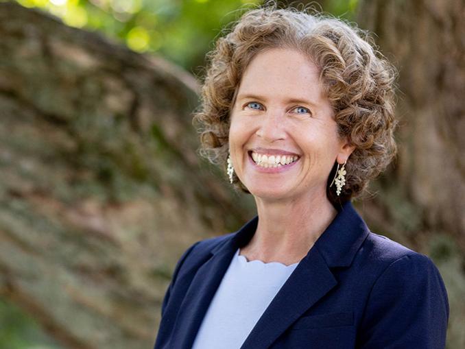 McDougall is pictured outdoors with trees in the background. She is smiling and wearing a light blue shirt and navy blue blazer.