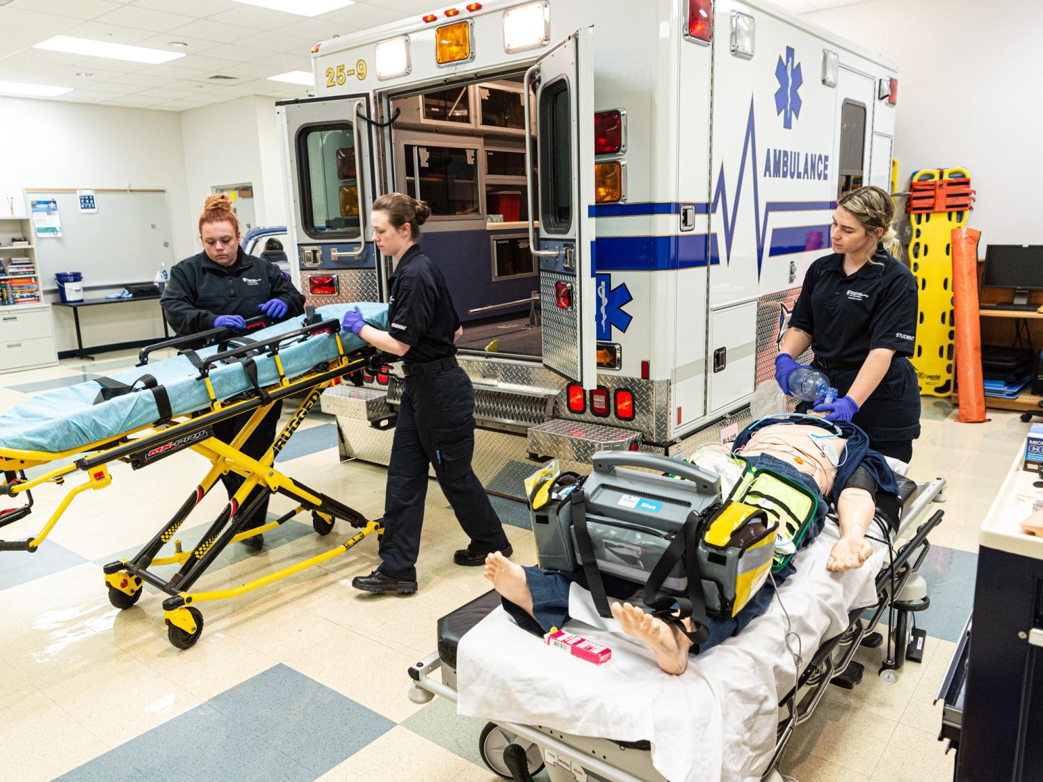 Student paramedics work with stretchers. An ambulance is behind them.