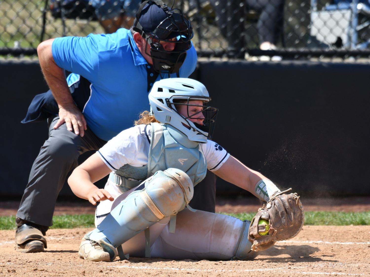 A catcher for the Penn State Behrend softball team grabs the ball as the umpire leans over her shoulder.