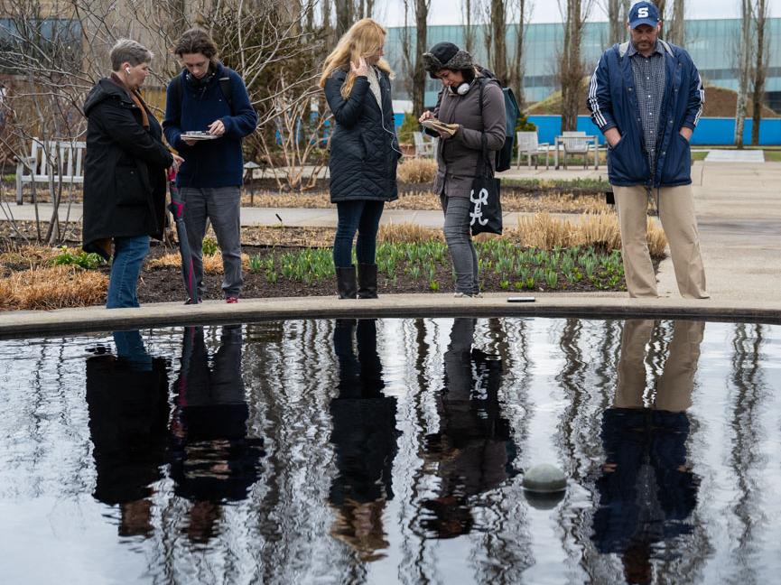A group of men and women, each holding a cell phone, stand in front of a reflecting pond.