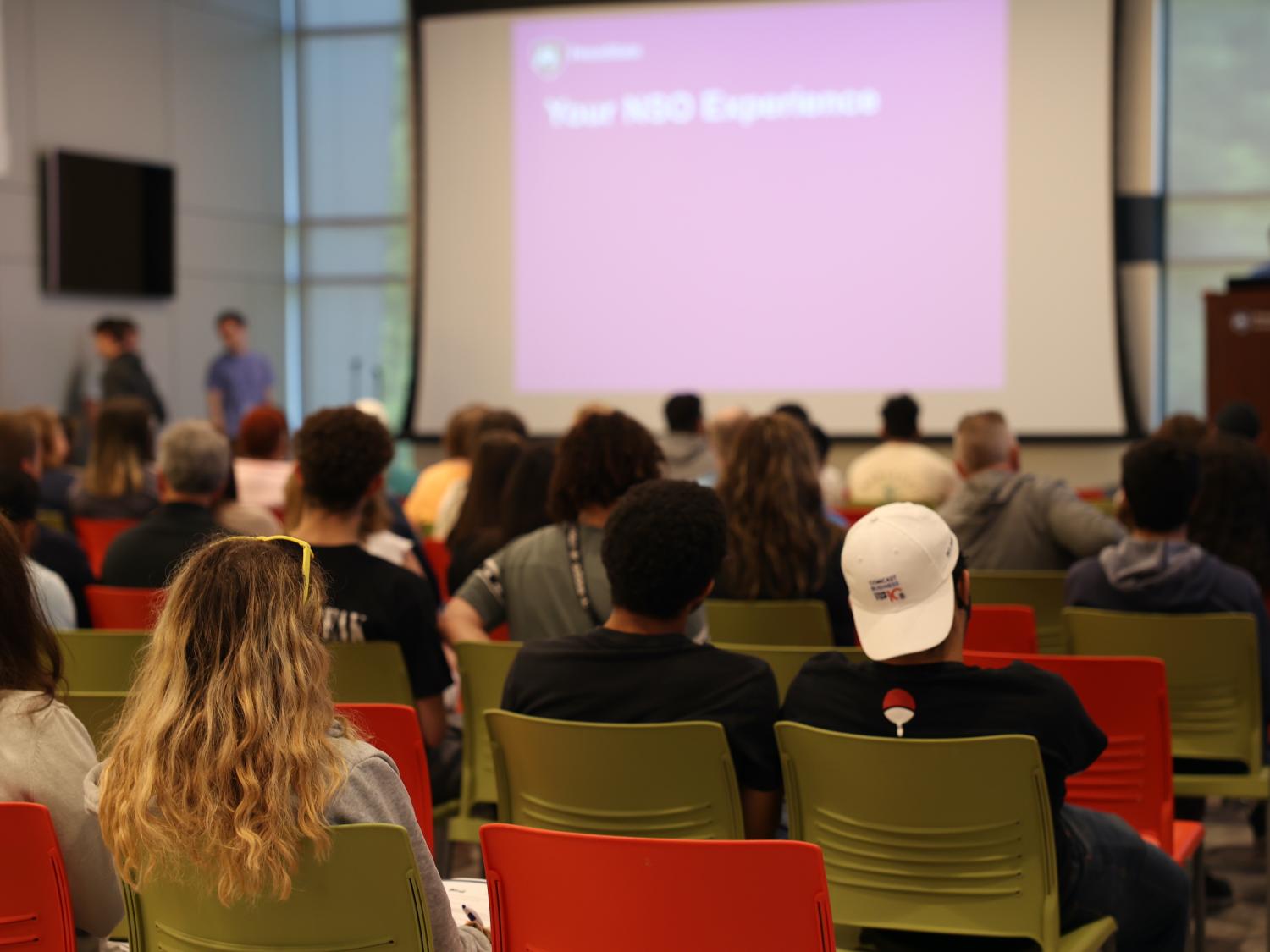 group of people sitting in chairs facing podium and screen