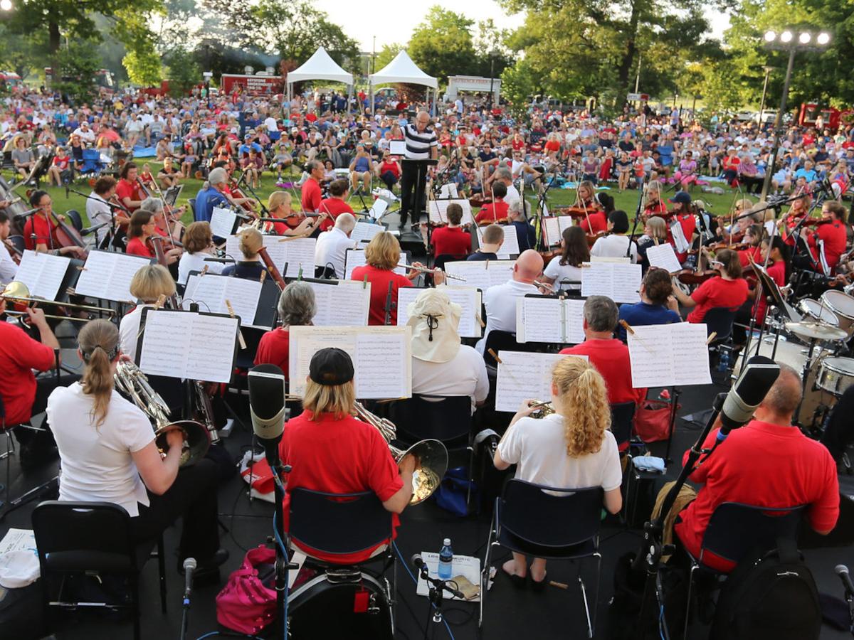 View from the back of an orchestra full of musicians performing before a crowd of a couple thousand people in an outdoor setting.