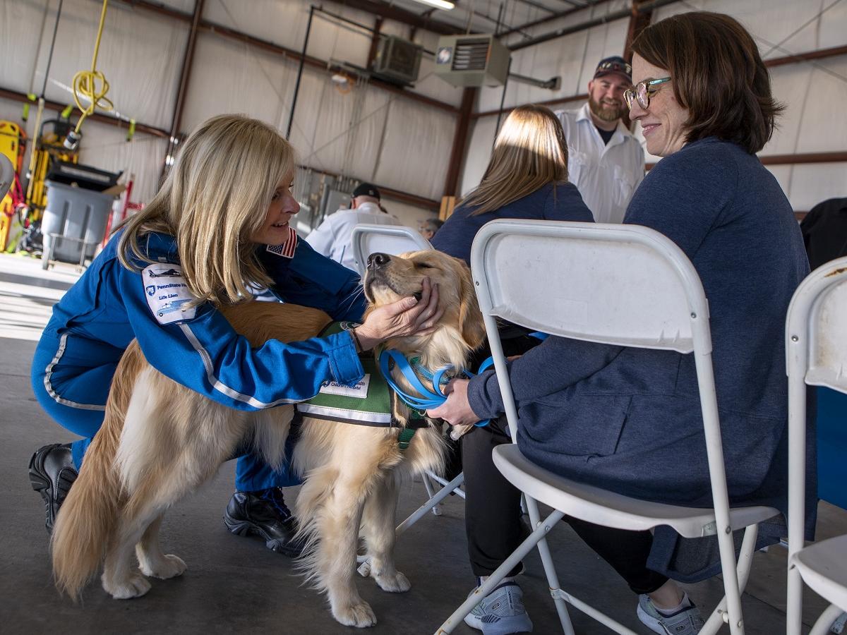 A woman pets a dog next to a woman seated in a chair.
