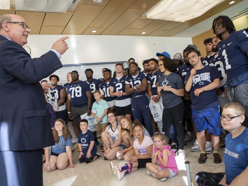 A man in a suit smiles and gestures at football players in their jerseys and a group of children.