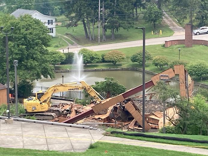 A piece of construction equipment sits in front of the remnants of a building. There is a pond with a fountain in the background.
