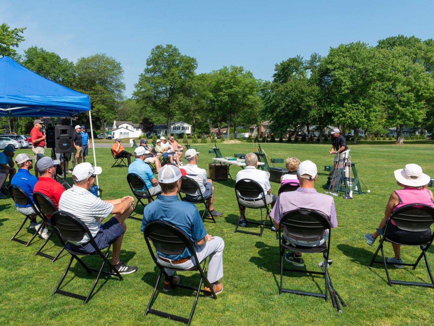 A group of people sit in lawn chairs and watch an exhibition given by a golf pro. It's a beautiful, sunny day.