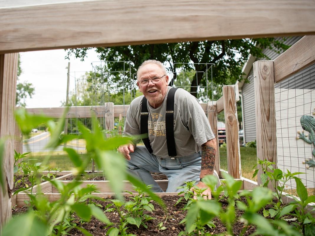 John Kline, wearing suspenders and glasses, kneels on the ground as he looks over his garden. Plants are visible in wooden planter beds. In the background are trees and a building on the right. In the far-right side of the photo, a decorative frog hangs on the garden door.