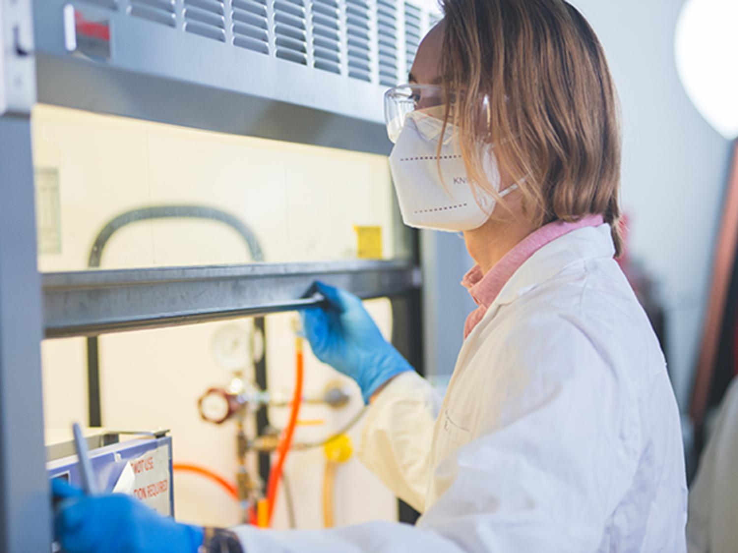 A person in personal protective equipment sits in front of a fume hood