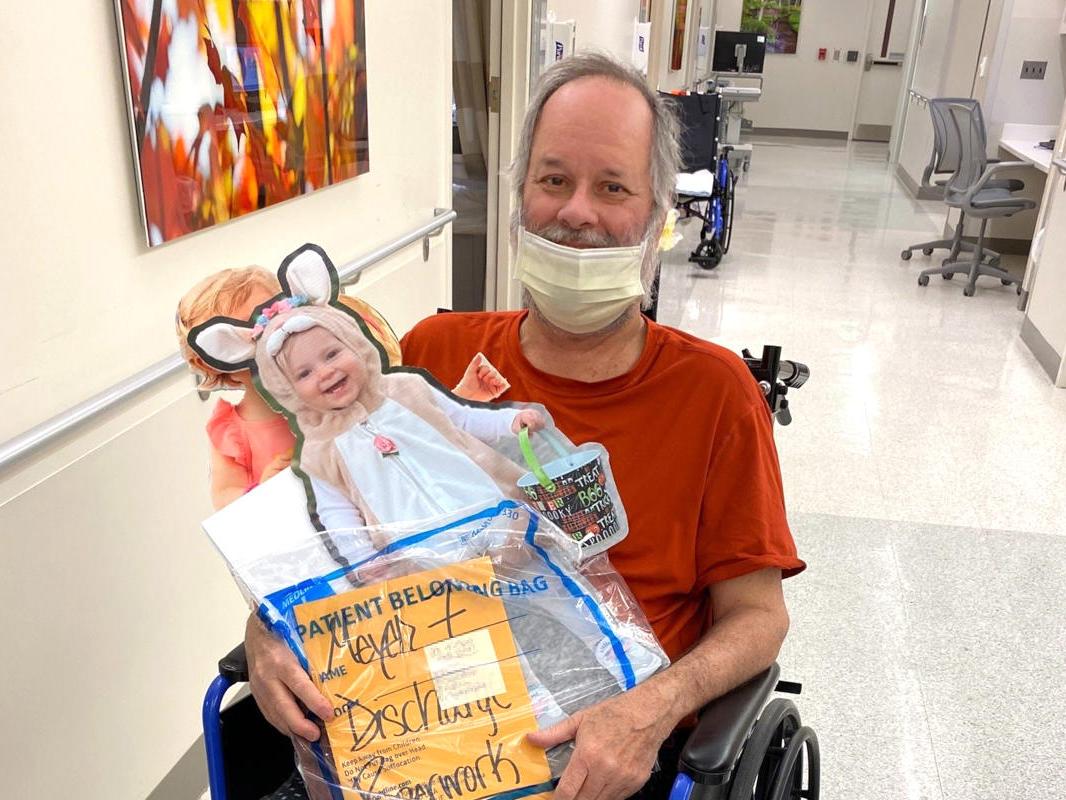 Man wearing a medical mask, seated in a wheel chair, holds an enlarged cardboard cutout of a photo of a little girl in an Easter costume and his hospital discharge paperwork.