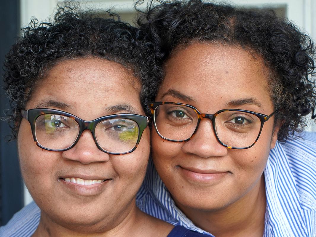 Twin sisters Alana and Alicia Webb smile as they pose in front of a window at their home. Both women have short curly hair and wear glasses and blue shirts.