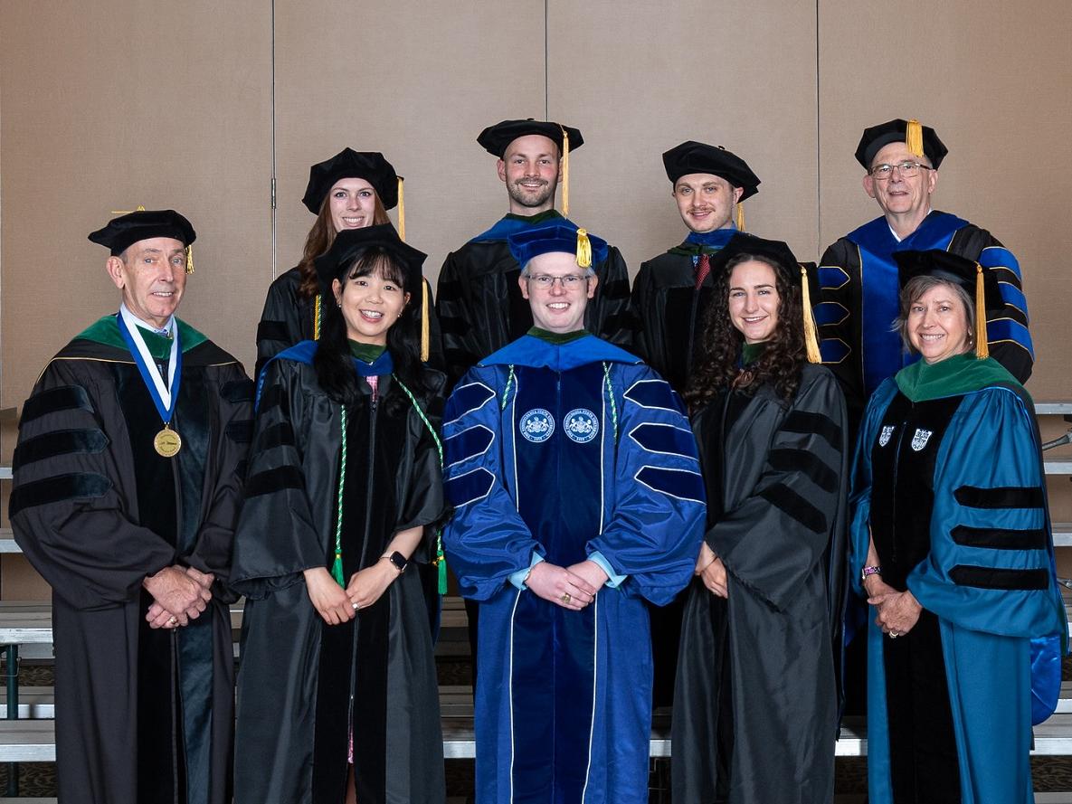 Nine people pose for a picture while wearing their academic regalia. 