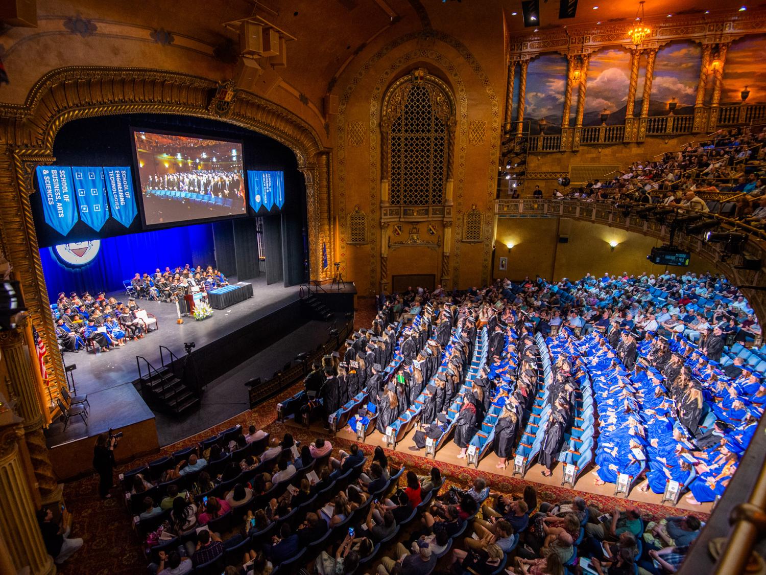 An auditorium filled with graduating students wearing caps and gowns listening to speakers on the stage