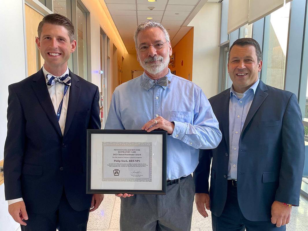 Three men stand in a row. One in the center holds a plaque.