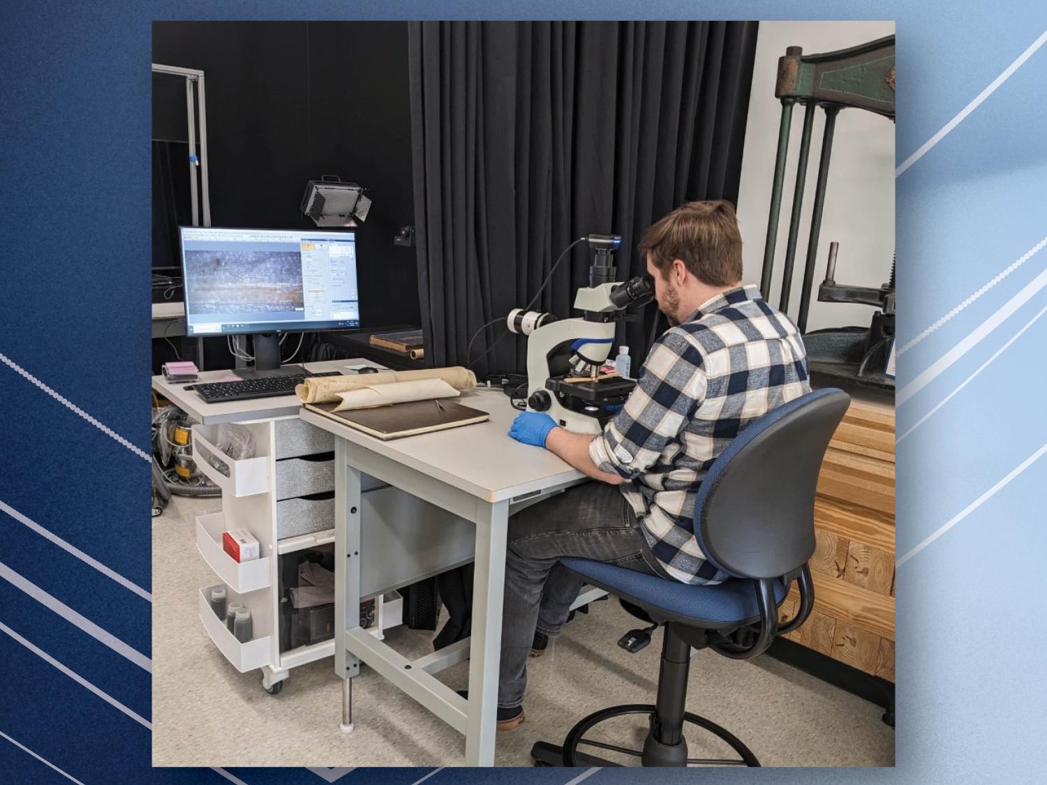 person sitting at table looking into microscope with computer monitor displaying what man is seeing in microscope