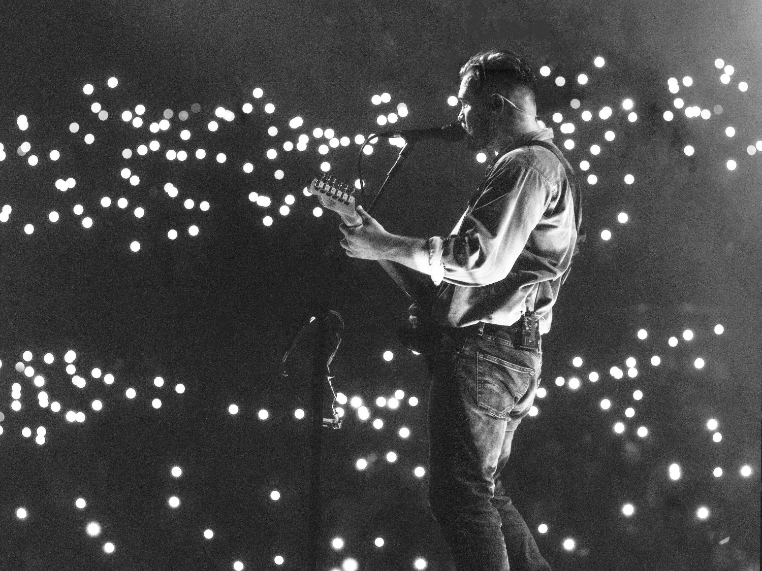 Zach Bryan on stage with a guitar and lights all around him