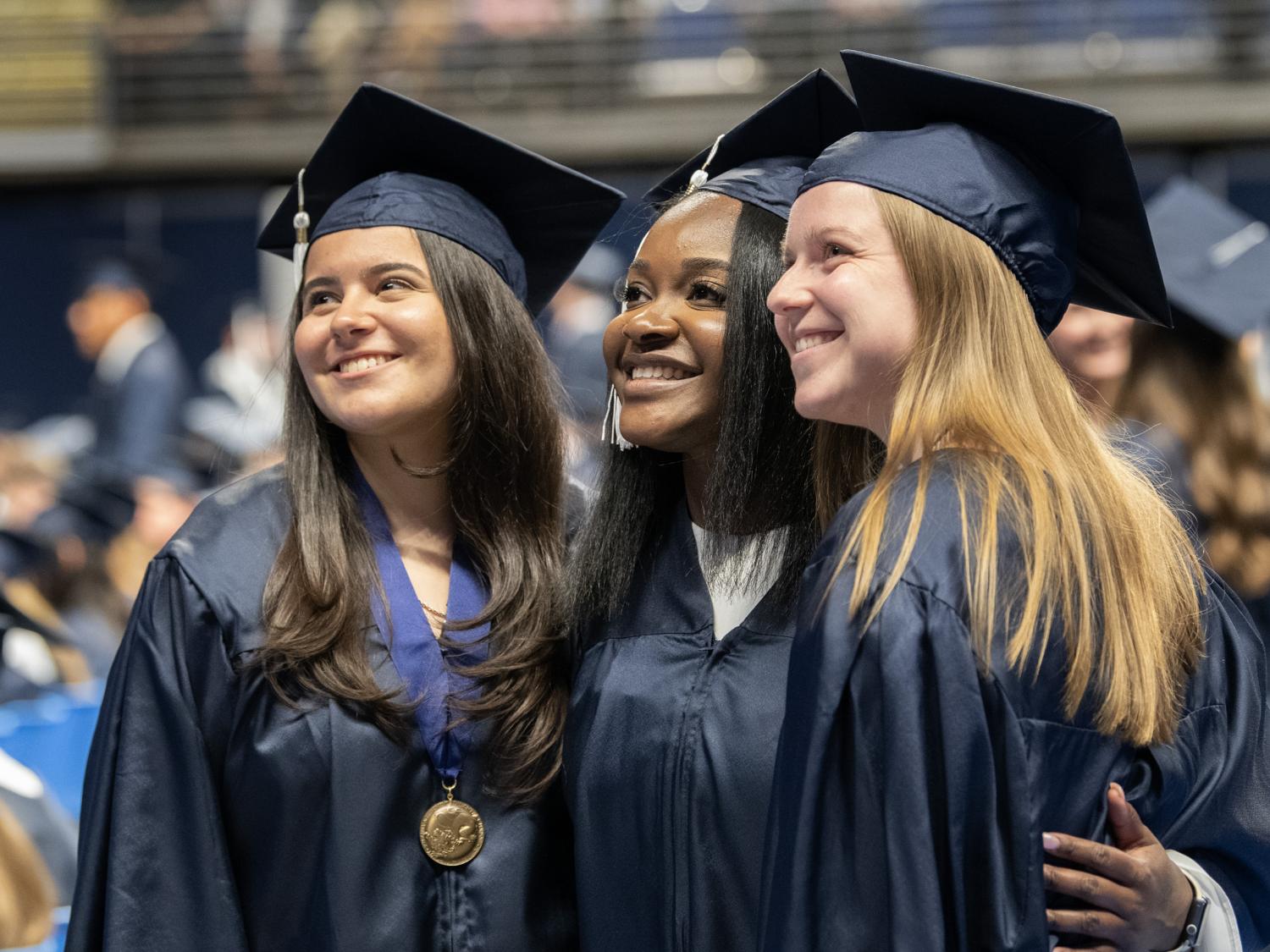 Three smiling women pose for a photograph, wearing blue graduation caps and gowns