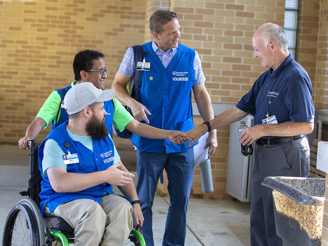Three men in blue hospital volunteer vests, one in a wheelchair, talk to a male hospital employee in a hospital entryway.