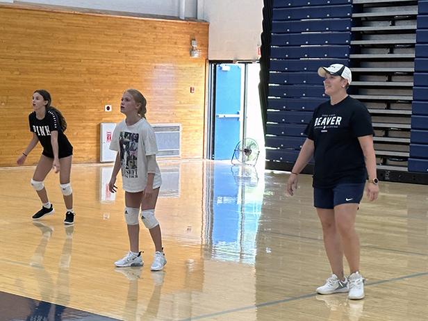 A female coach wearing navy blue and a ball cap stands to the right of the photo in a gymnasium as female volleyball players prepare to make a play to her left.