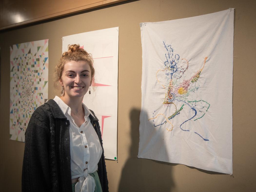Abigail Wallar standing next to her embroidered design that is hanging on a display board. 