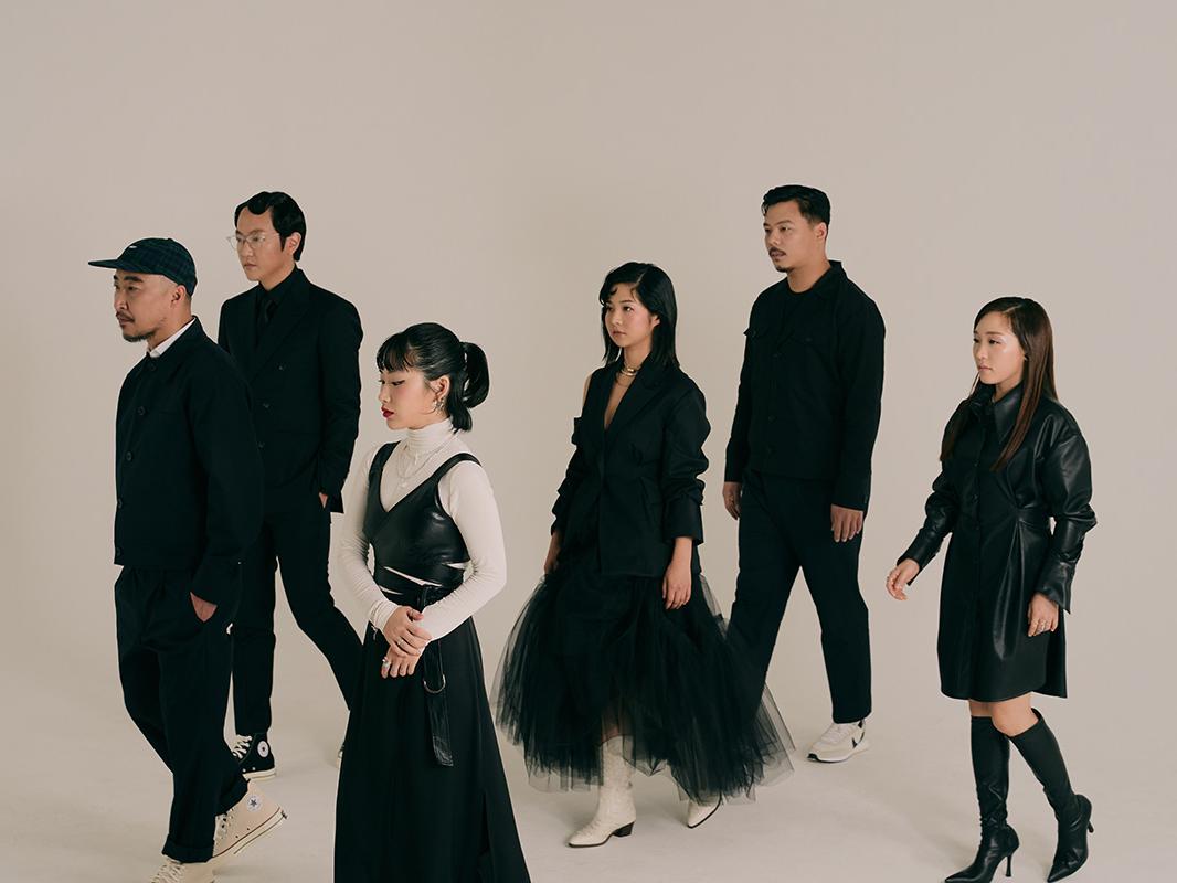 A group of six Korean musicians wearing monochromatic outfits stand and look in the same direction.