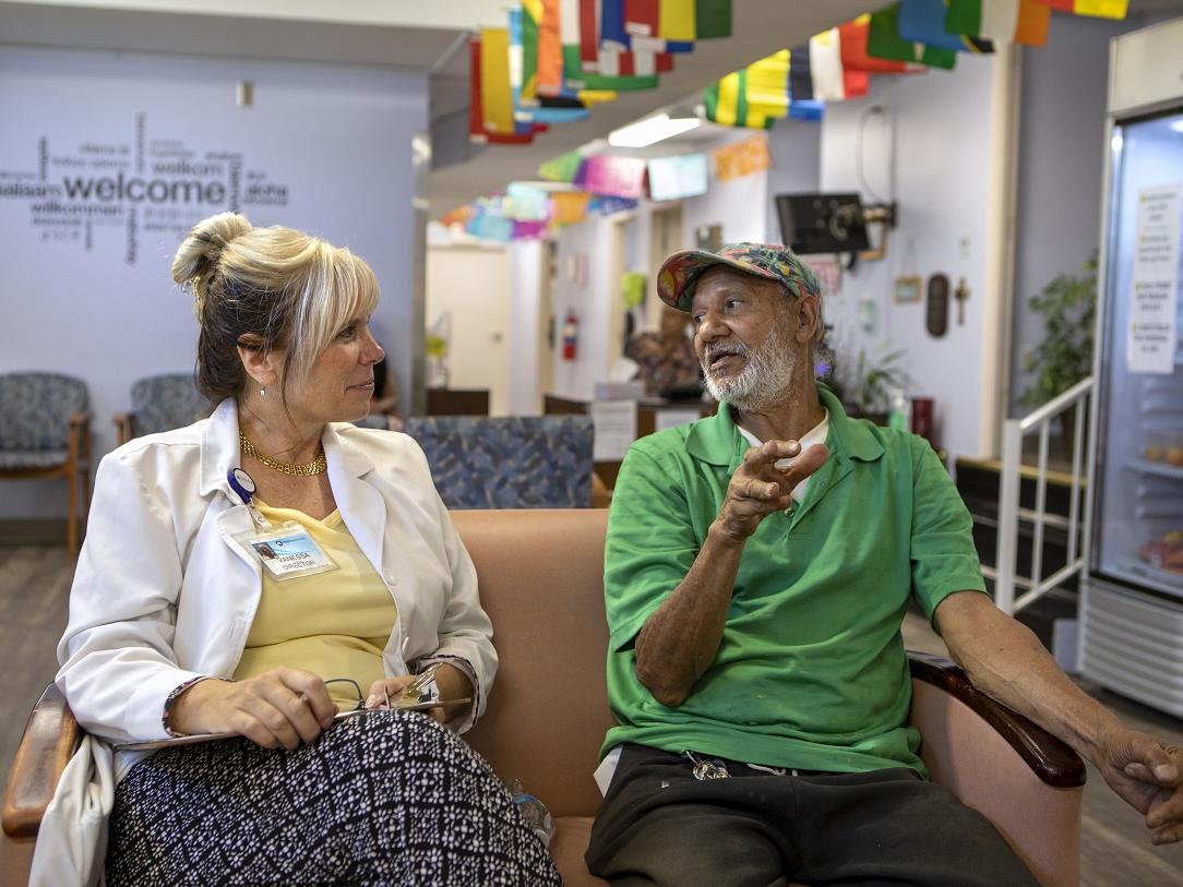 A man and a woman sit side by side on a couch in a waiting room. Overhead, flags from many different countries are attached to the ceiling.