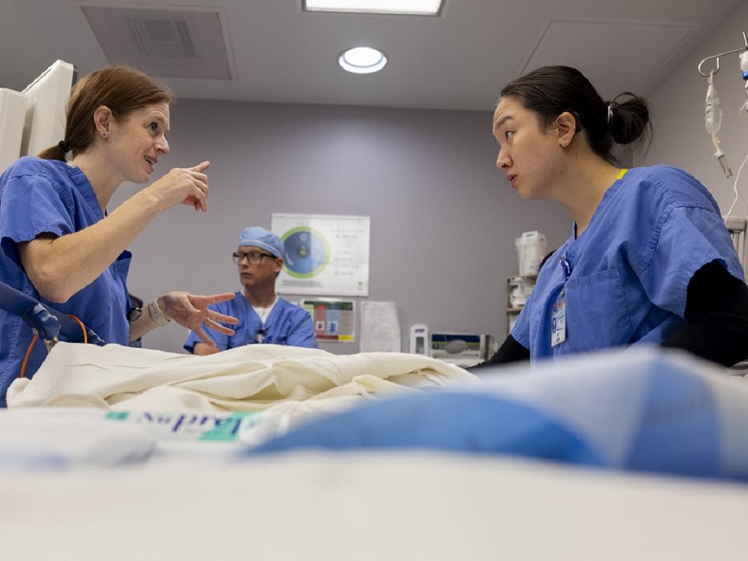Two women lean across a hospital bed. The woman on the left gestures at the woman on the right. A man in the background looks on.