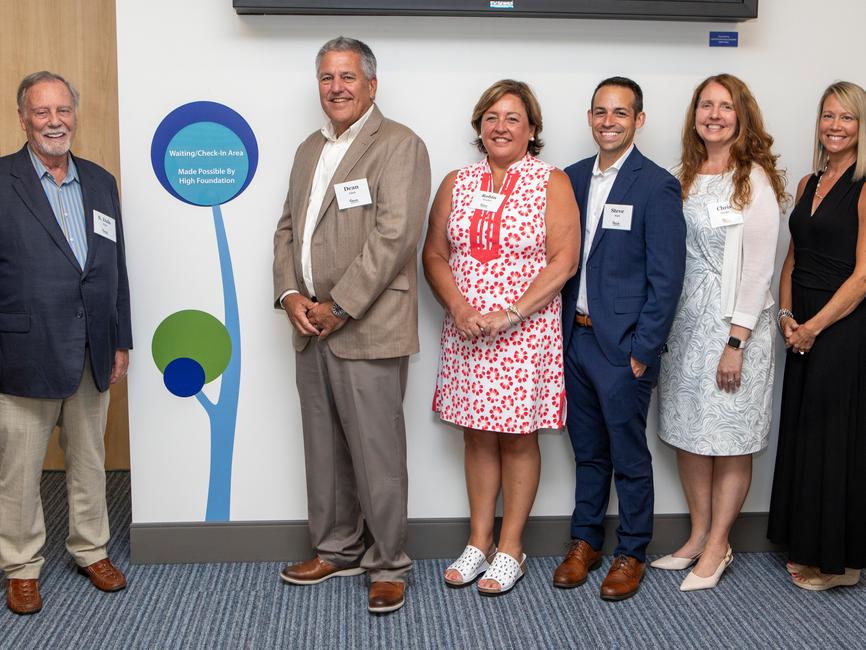 Six people wearing professional attire pose for a photo. In the middle is a sign on a wall reading: “Waiting/Check-In Area Made Possible By High Foundation.”