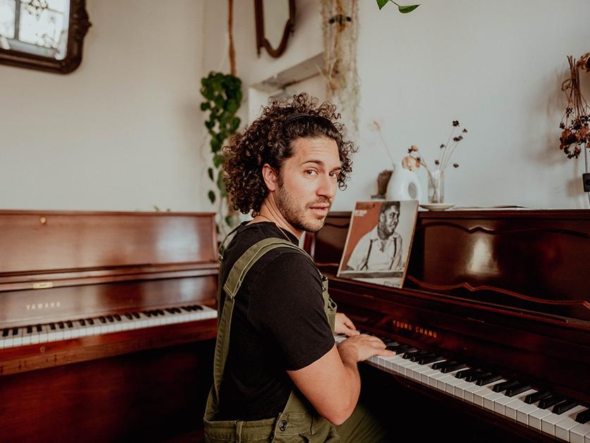 A man sitting at the corner of two piano keyboards looks at the camera.