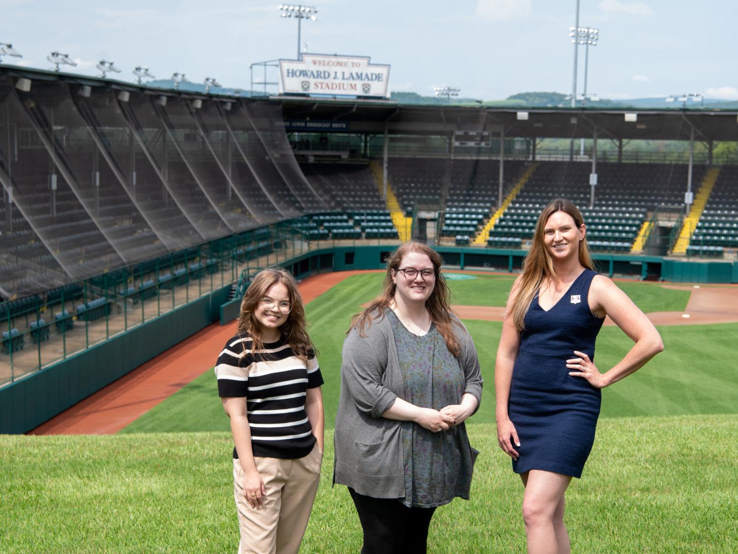 Three people stand on a softball field with the stands in the background