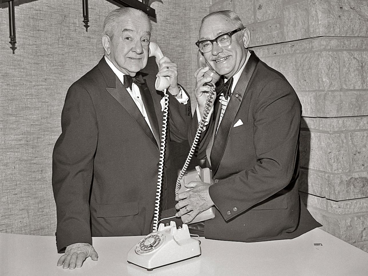Two smiling men in suits hold phone receivers to their ears in this black and white historical photograph