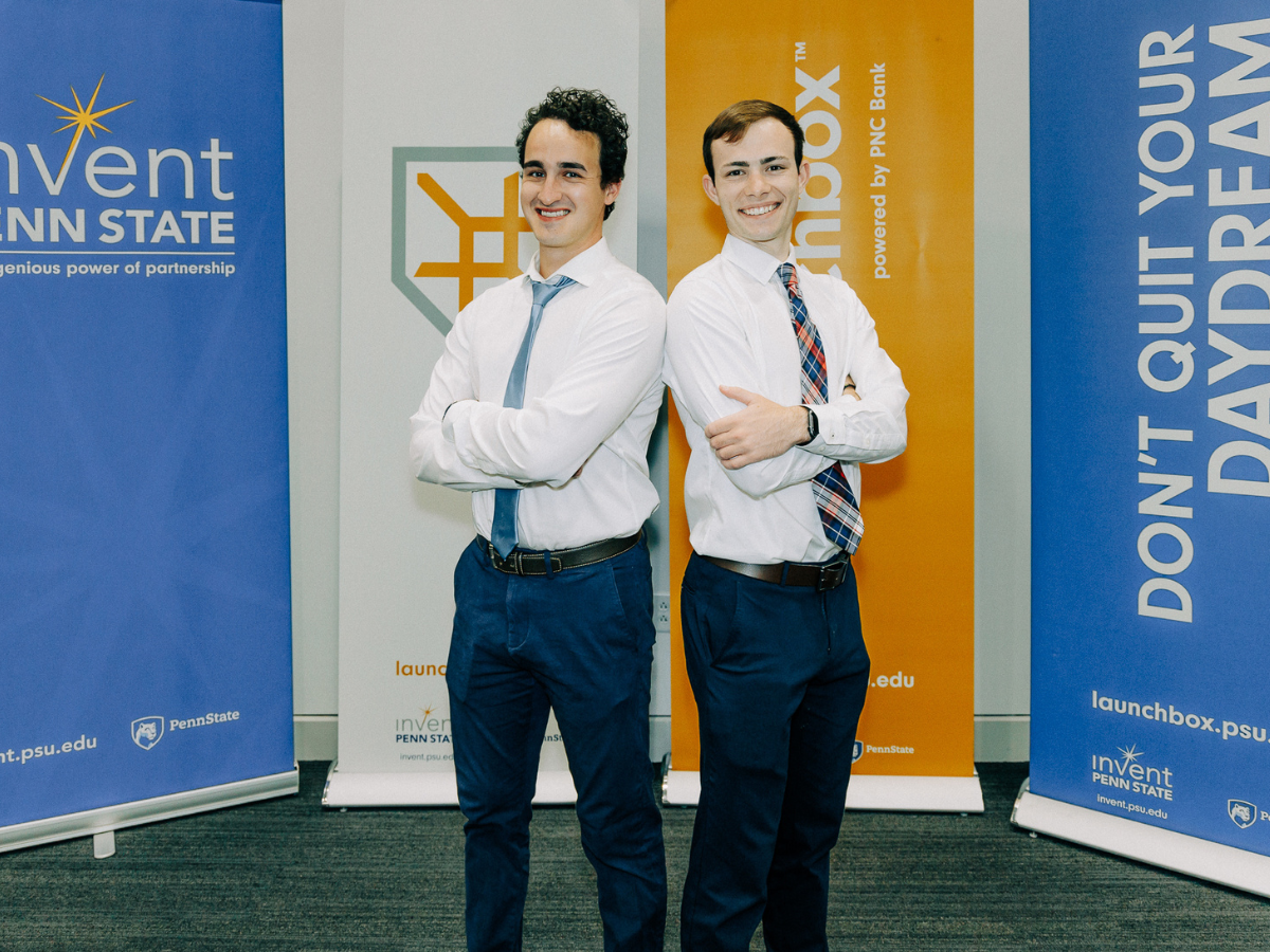 Business partners and Penn State students Noah Wiggins and Nolan Pye pose for a photo in front of Invent Penn State signage