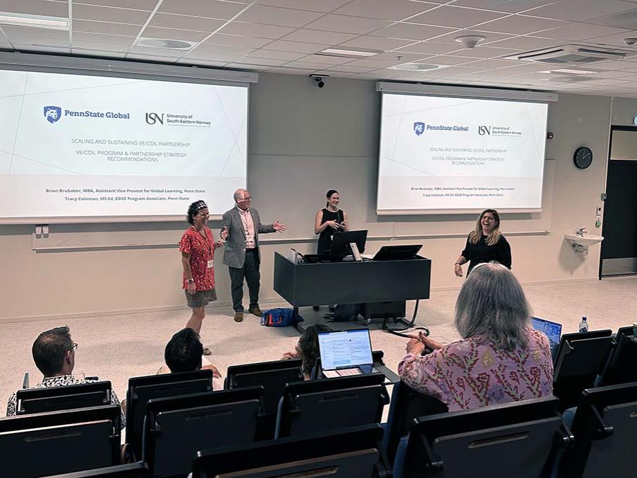 Four people standing in front of an audience in a classroom with two video projector screens on either side of them.