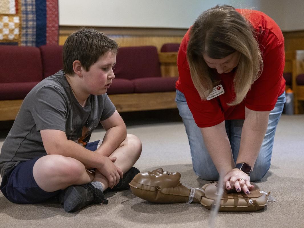 A young boy watches a woman perform chest compressions on a medical doll.