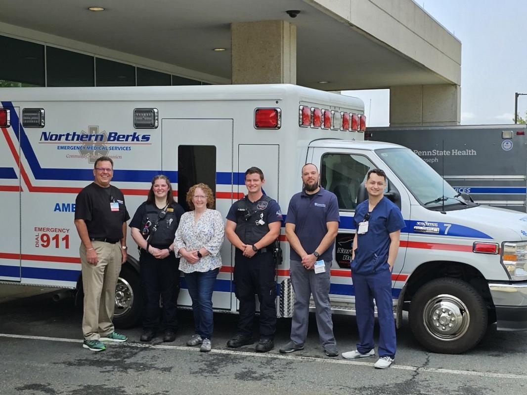 Six adults stand in front of an ambulance parked in front of a hospital. A second ambulance is visible behind it to the right. The man standing on the left and the two men on the right have name badges clipped to their clothing. The woman second from left and the man fourth from left are wearing vests outfitted with a radio/intercom system. The woman has sunglasses clipped to her vest.