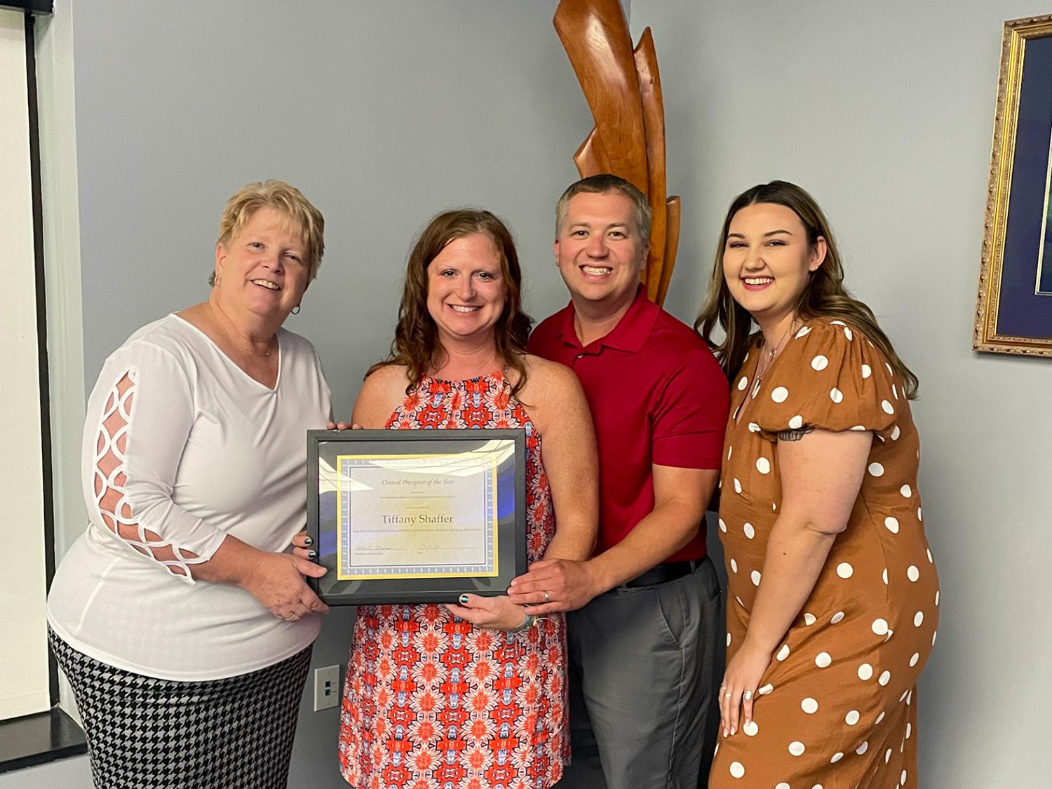 Four people pose for a photograph with the center person holding a framed certificate.
